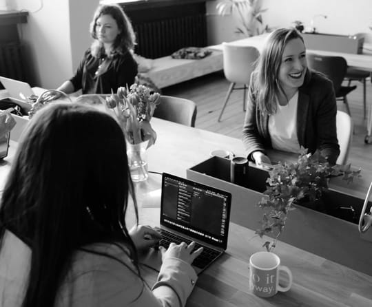 Three woman sitting in an office, two of them are writting something on their laptops, the third one is smiling