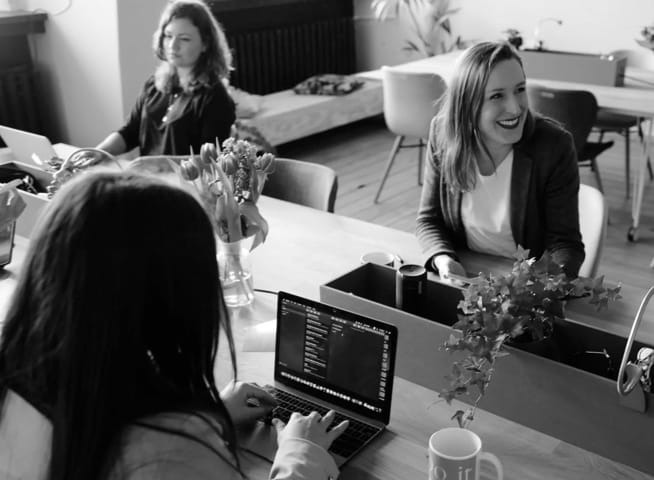 Three woman sitting in an office, two of them are writting something on their laptops, the third one is smiling